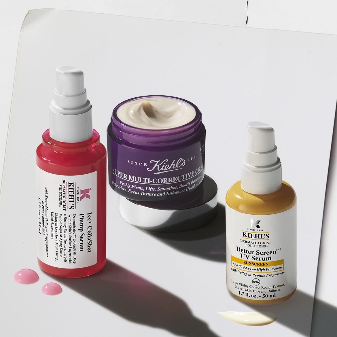 Kiehl's 1cc CollaShot Plump Serum in een roze flesje, Super Multi-Corrective Cream in een paarse pot en Better Screen UV Serum in een gele flesje, met productdruppels op een witte achtergrond.
