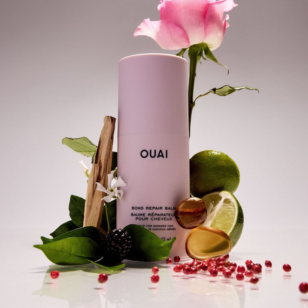Le baume réparateur de OUAI, présenté dans un emballage rose pâle, est entouré d'ingrédients naturels comme une rose, des citrons verts, des baies et des feuilles, évoquant une formule fraîche et nourrissante pour les cheveux abîmés.
