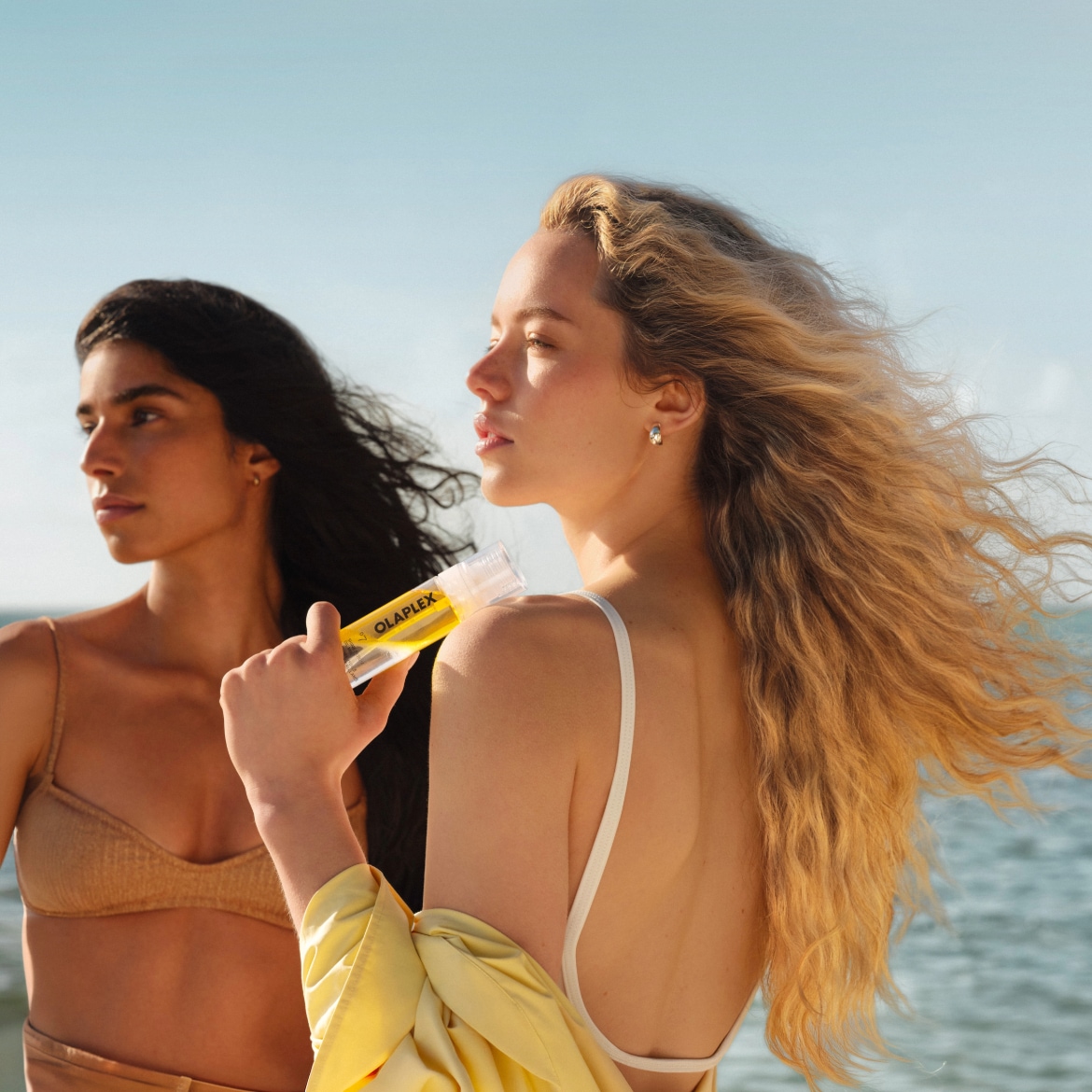 Twee vrouwen staan bij de oceaan, de ene met donker haar en de andere met blond haar, die een Olaplex-product vasthoudt.