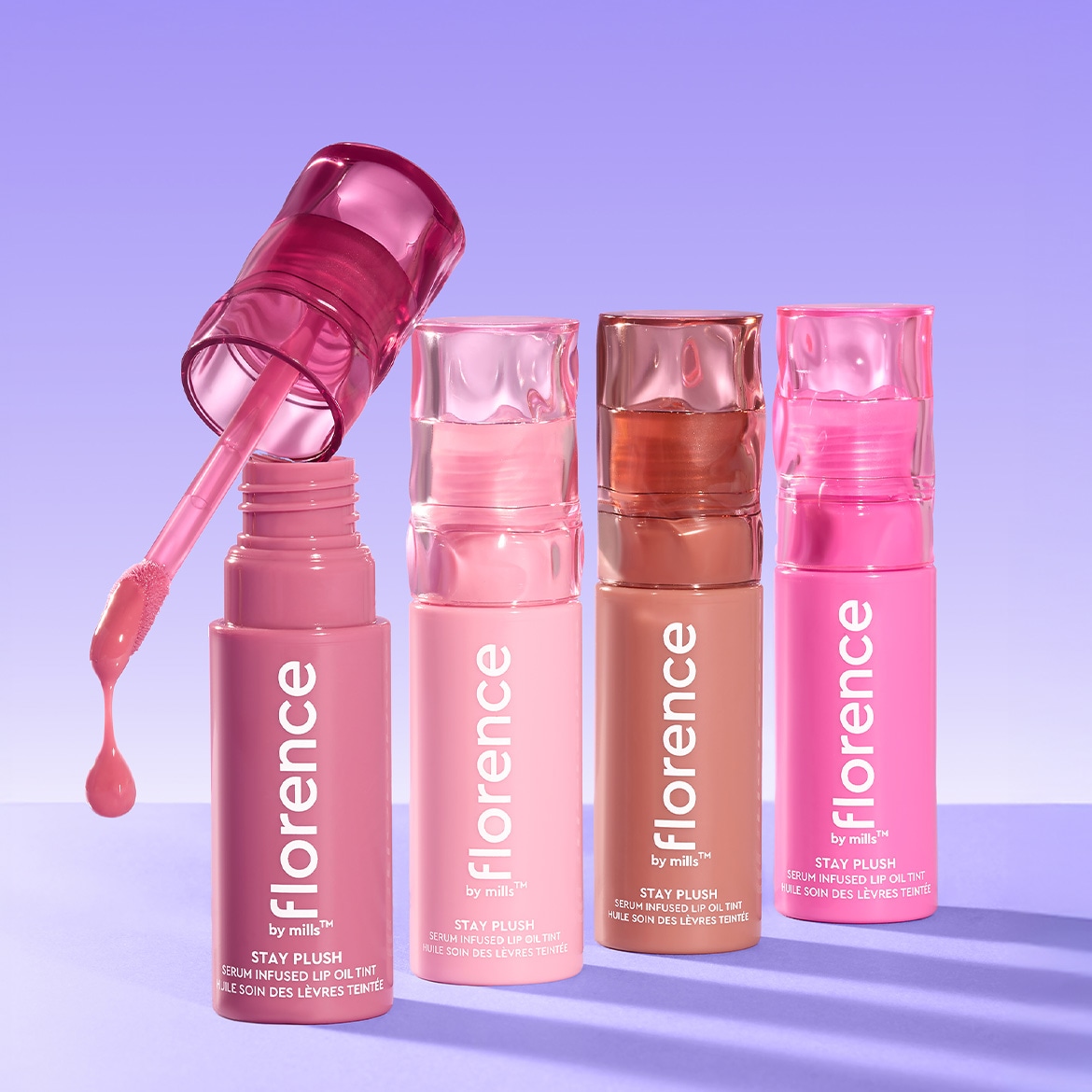 Vier Florence by Mills Stay Plush Serum Infused Lip Oil Tint flacons in verschillende roze en bruine tinten, met één open flacon waaruit een roze lipolie druppelt.