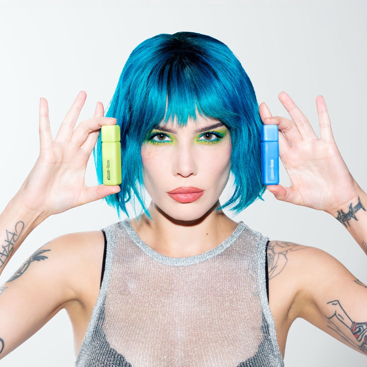 Une femme aux cheveux bleus vibrants présente deux produits de beauté : un flacon vert clair et un flacon bleu, chacun promettant une peau éclatante et fraîche.