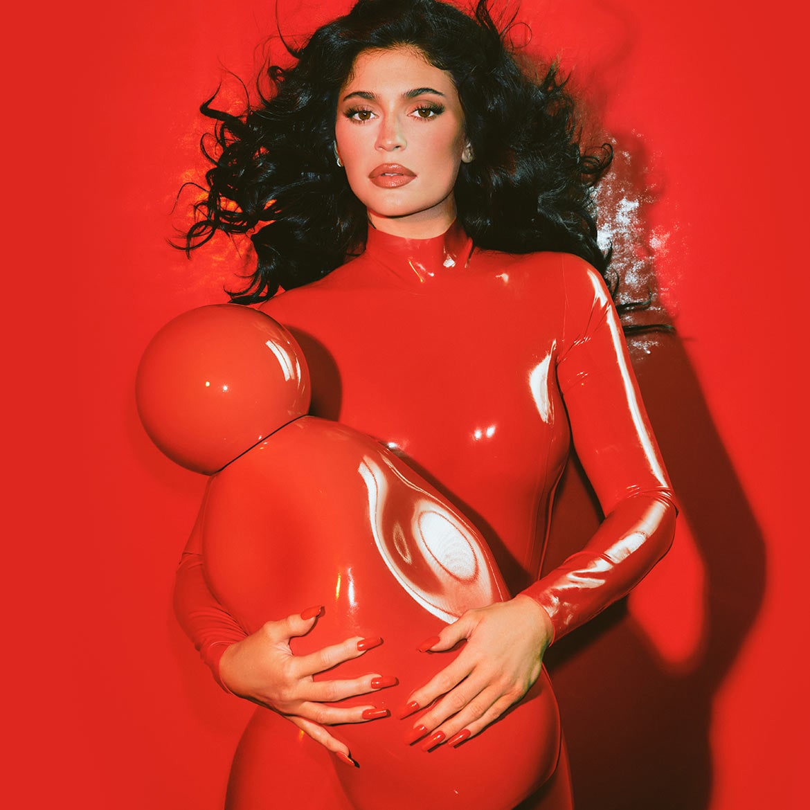 Kylie Jenner draagt een rode latex bodysuit en houdt een rode, bolvormige prop vast tegen een rode achtergrond.