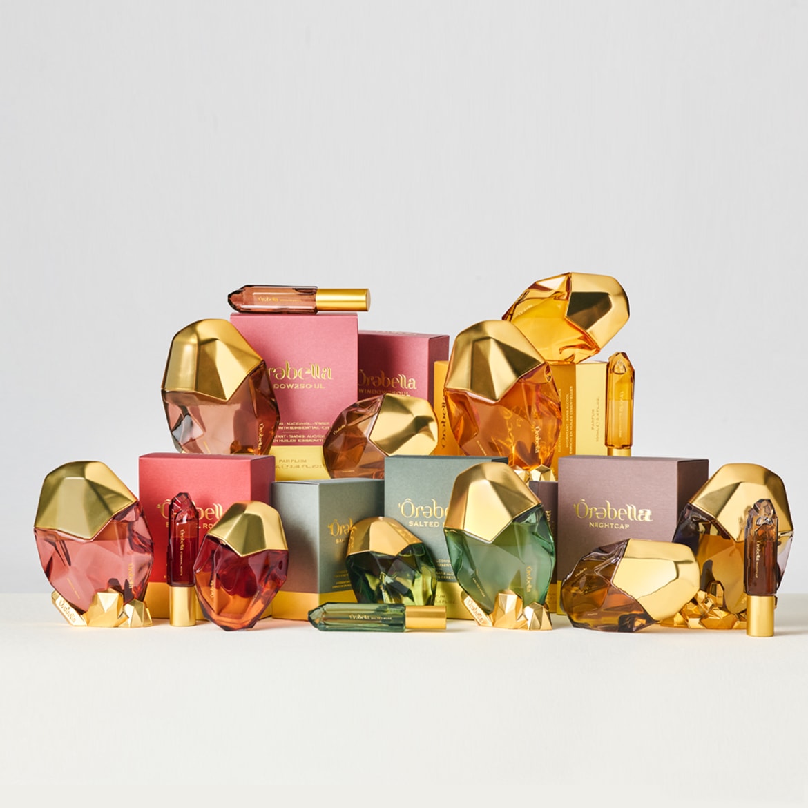 Ontdek de luxe collectie van Òrabella parfums, gepresenteerd in veelkleurige flacons met gouden accenten en bijpassende verpakkingen.