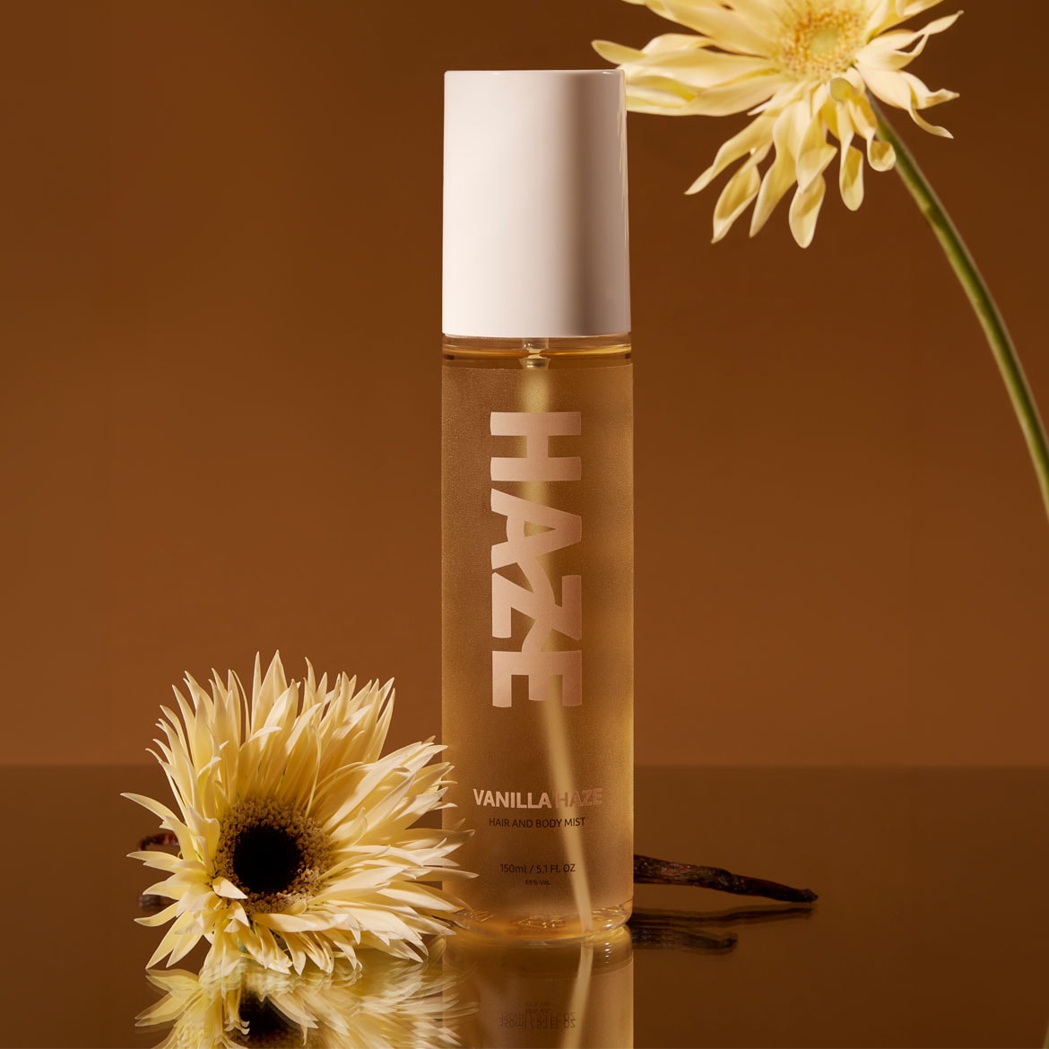 De Vanilla Haze Hair and Body Mist zit in een doorzichtige fles met een witte dop, versierd met gele bloemen en een vanillestokje.