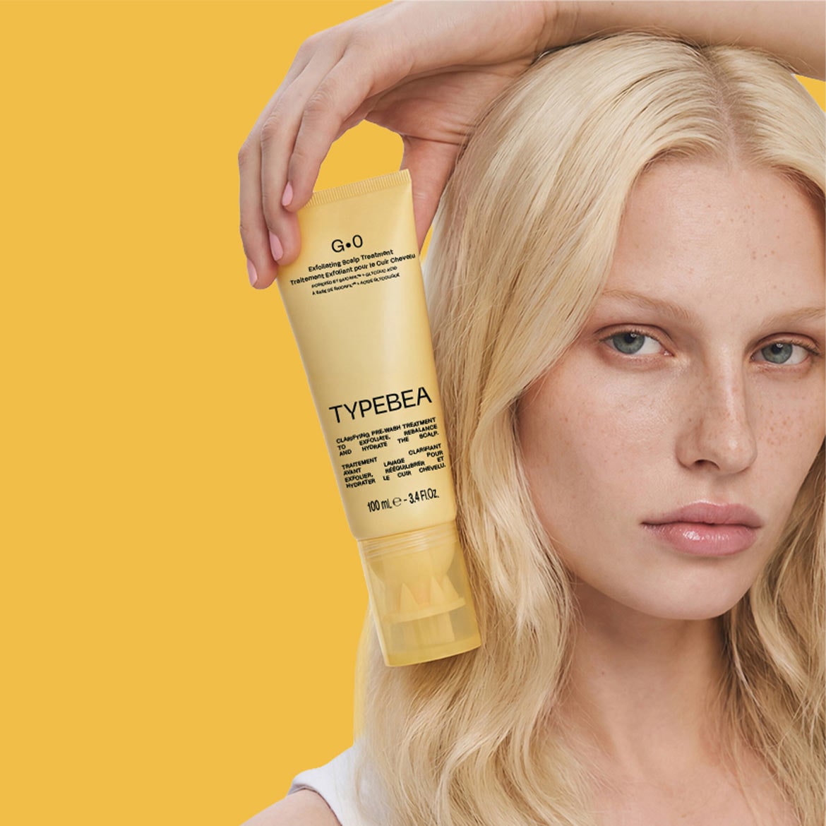 Een vrouw met blond haar houdt een gele tube G.O. Exfoliating Scalp Treatment van Typebea vast, een zuiverende pre-wash behandeling die de hoofdhuid exfolieert, hydrateert en in balans brengt.