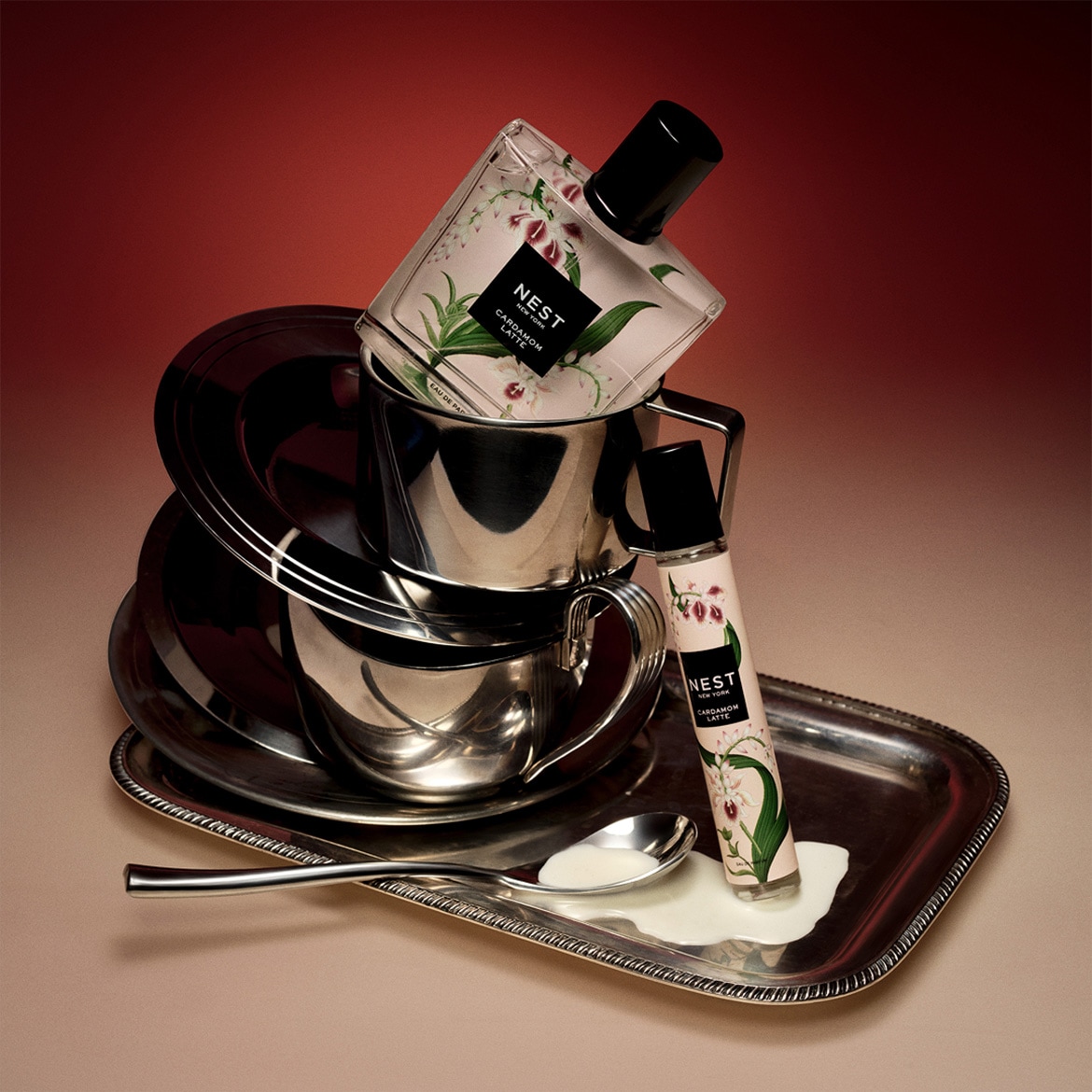Le Nest New York Cardamom Latte Eau de Parfum et un rollerball sont posés sur un plateau en argent avec une cuillère et du lait renversé, entourés de tasses et soucoupes en argent.