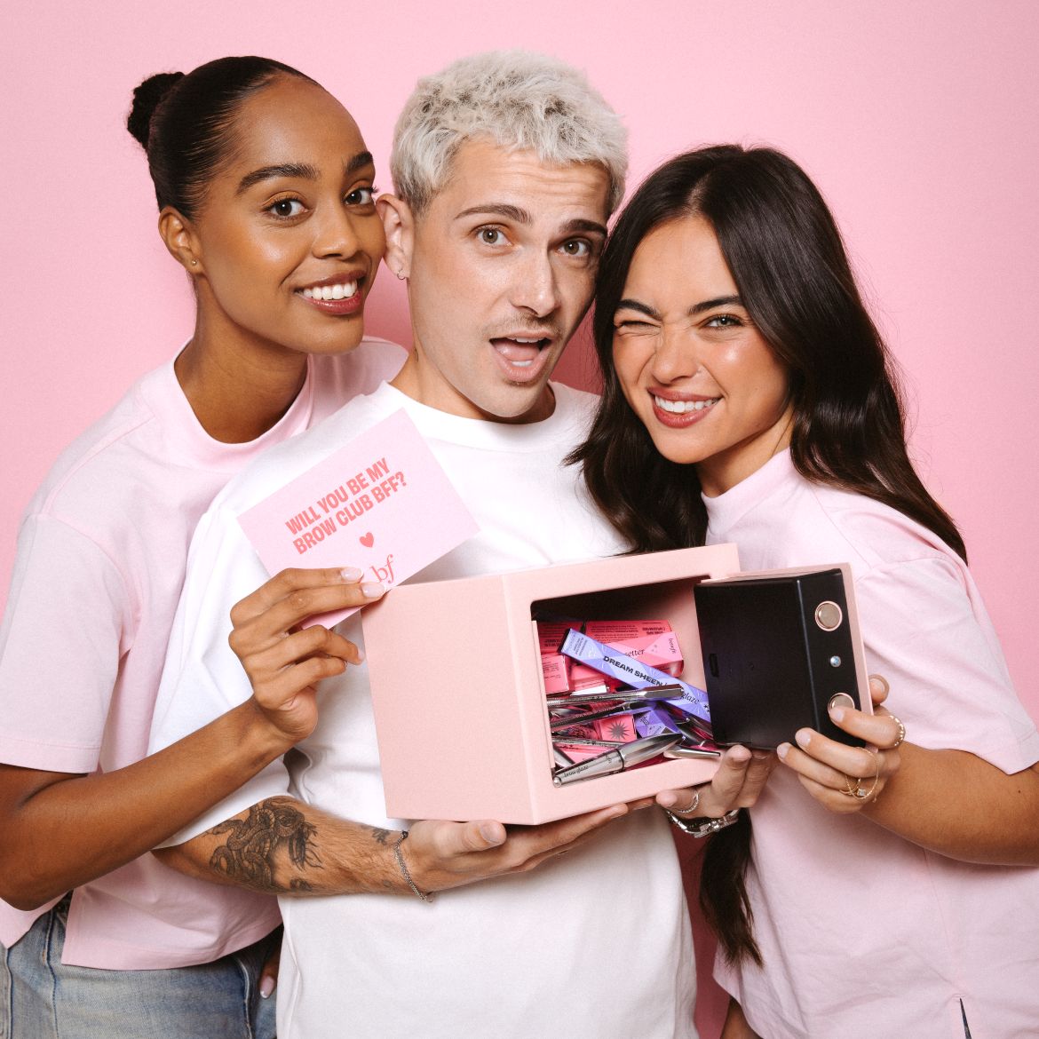 Trois personnes souriantes posent avec un coffre-fort rose rempli de produits pour les sourcils Benefit et une carte "WILL YOU BE MY BROW CLUB BFF?".