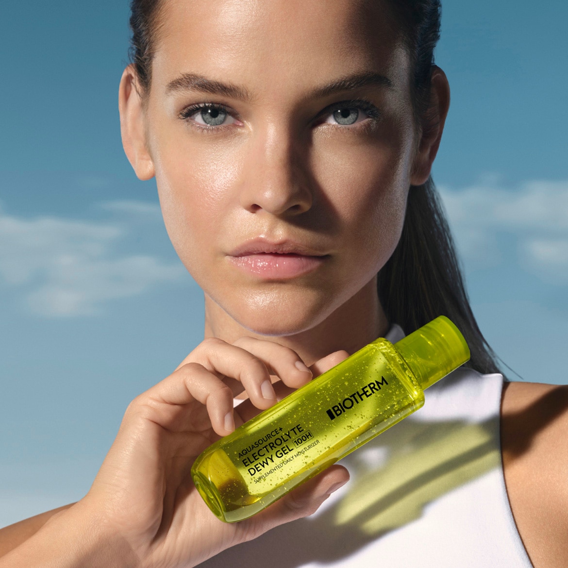 Een vrouw houdt een heldere, geelgroene fles Biotherm Aquasource Electrolyte Dewy Gel 100H vast tegen een blauwe lucht, wat de hydraterende en verfrissende eigenschappen van het product benadrukt.