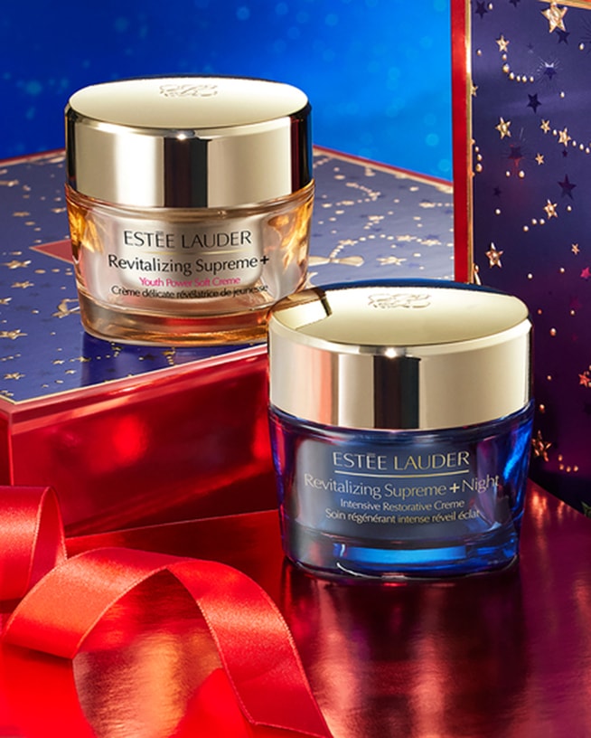 Twee potten Estée Lauder Revitalizing Supreme+ crème, één voor overdag en één voor 's nachts, met een gouden deksel en een rode strik.