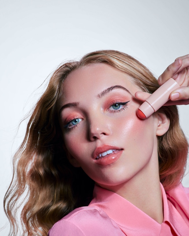 Een verzameling make-up producten, waaronder een rode lippenstift, Maybelline lipgloss, Pixi blush, NYX wenkbrauwlijm en Too Faced mascara, allemaal in zachte tinten.