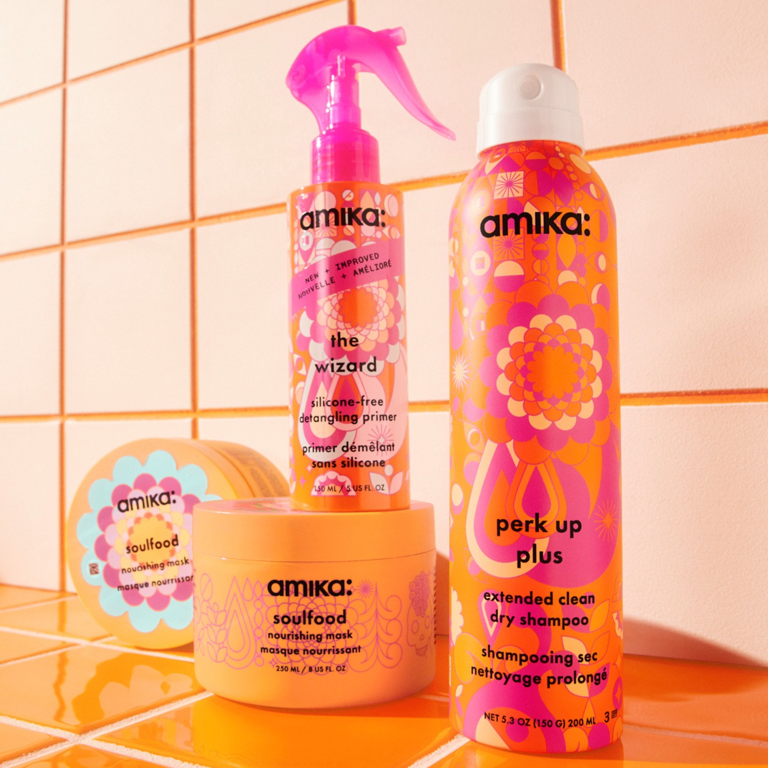Verschillende Amika haarverzorgingsproducten staan op een oranje tegelachtergrond, waaronder 'The Wizard' ontwarrende primer en 'Perk Up Plus' droogshampoo in felgekleurde verpakkingen.