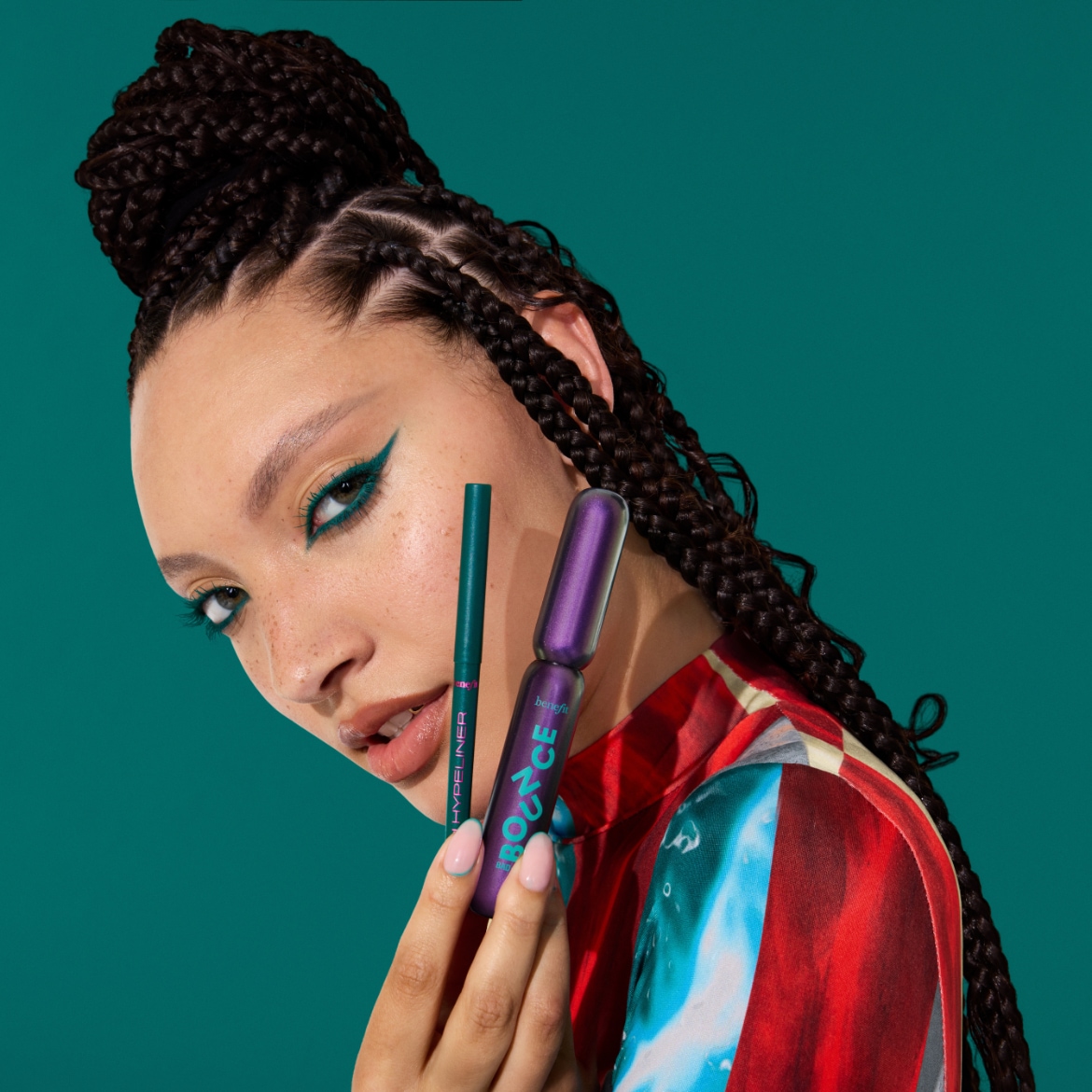 Een vrouw met gevlochten haar en een kleurrijke top houdt een groene eyeliner en een paarse mascara van Benefit Cosmetics vast, met een opvallende groene eyelinerlook.