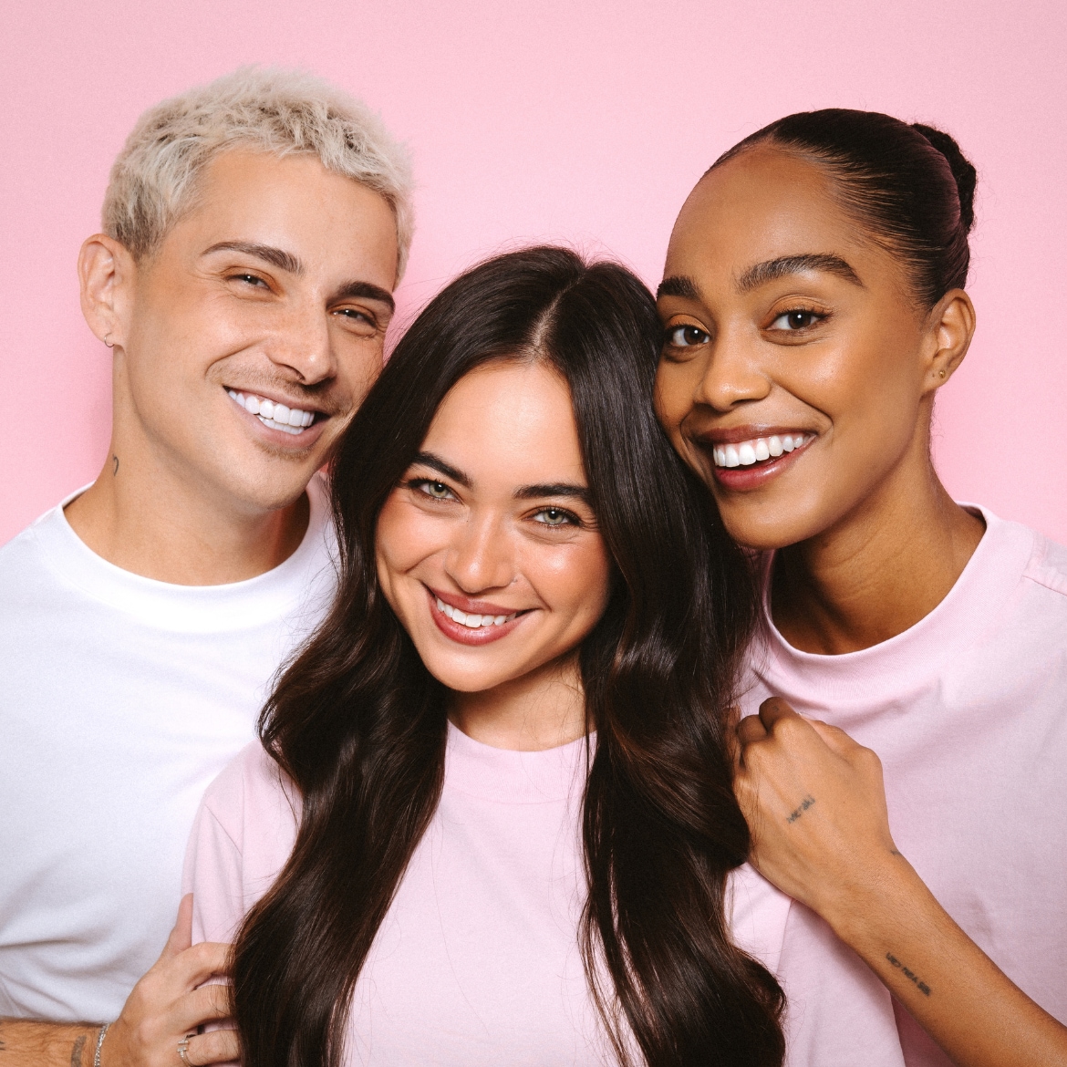 Drie lachende modellen met een stralende huid en witte tanden, twee dragen een roze T-shirt en één een wit T-shirt, tegen een roze achtergrond.