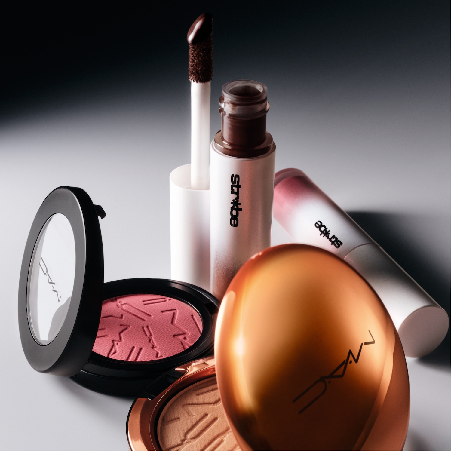 Een close-up van verschillende make-upproducten van MAC Cosmetics, waaronder een roze blush, een bronzer, een lipgloss en een vloeibare highlighter, allemaal elegant gerangschikt op een grijze achtergrond.