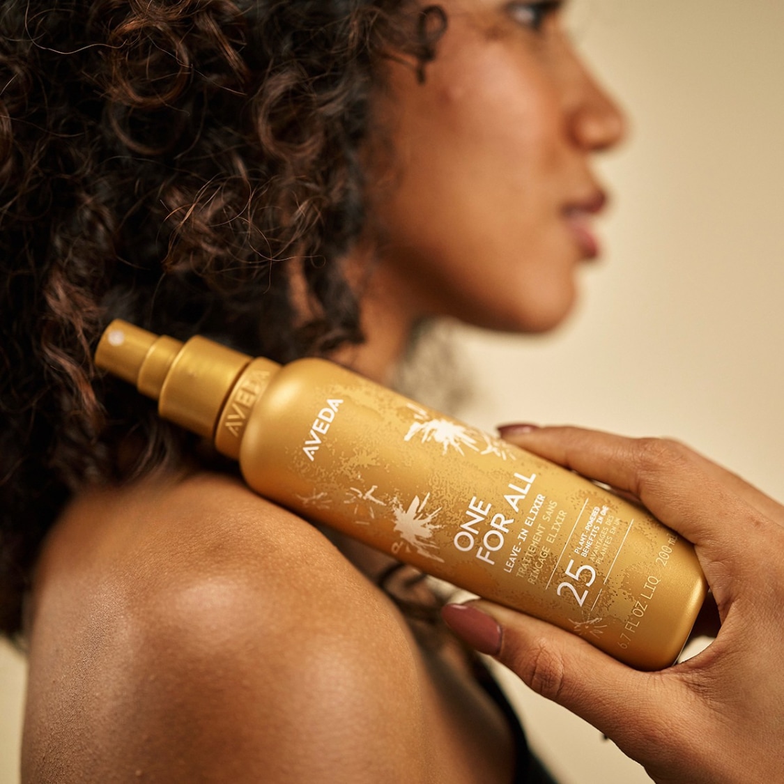 Een vrouw met krullend haar houdt een goudkleurige fles Aveda One For All Leave-In Elixir vast, met de tekst "25 plant-powered benefits in one" op de voorkant.