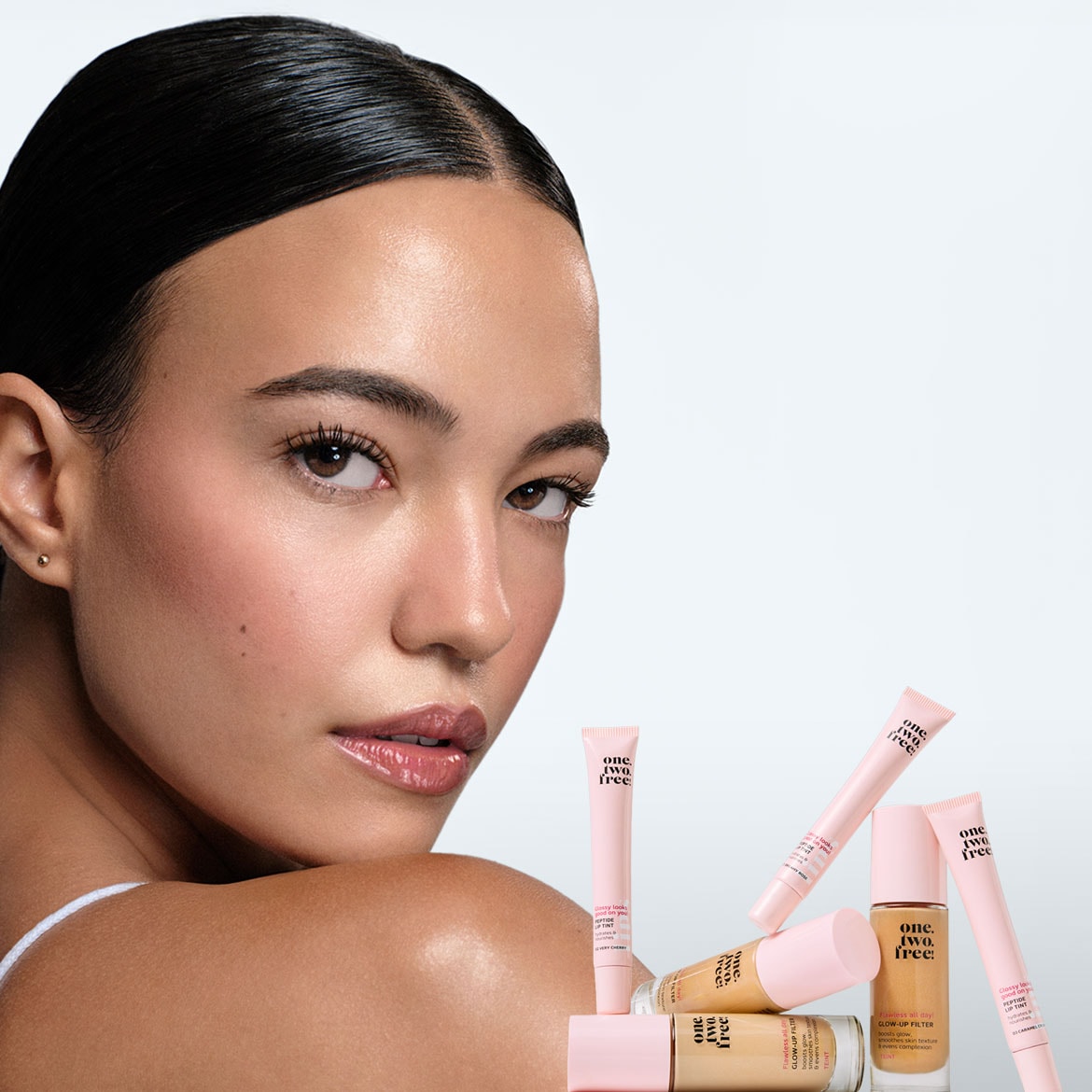 Une femme au teint éclatant et aux cheveux noirs lisses regarde par-dessus son épaule, avec plusieurs produits de maquillage One.Two.Free! aux tons nude et rose pastel disposés devant elle.
