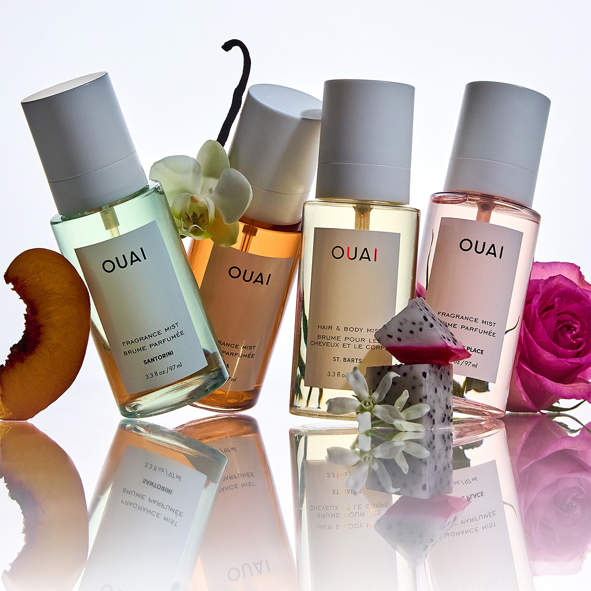 Vier flessen OUAI geurmist, elk met een unieke geur, variërend van Santorini tot St. Barts, gepresenteerd met complementaire bloemen en fruit.