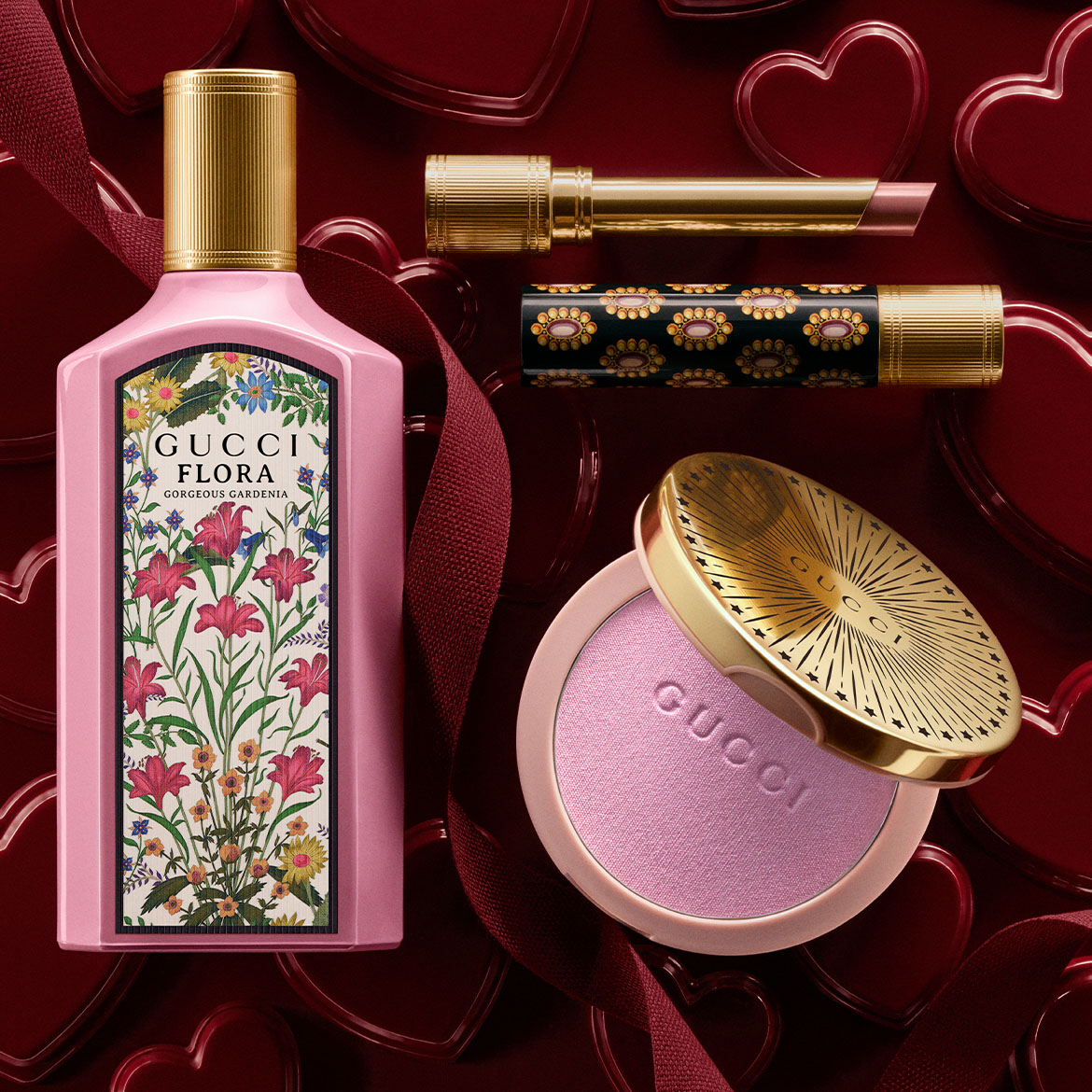 Découvrez le parfum Gucci Flora Gorgeous Gardenia dans un flacon rose, un rouge à lèvres doré et un blush rose poudré, le tout sur un fond rouge orné de cœurs.