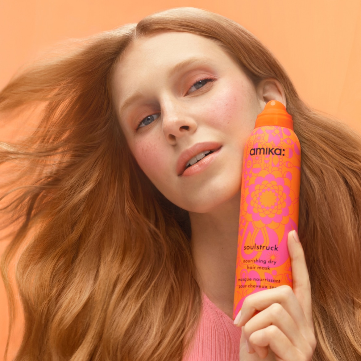 Une femme aux cheveux roux présente le masque capillaire sec nourrissant Amika Soulstruck dans un emballage orange et rose.