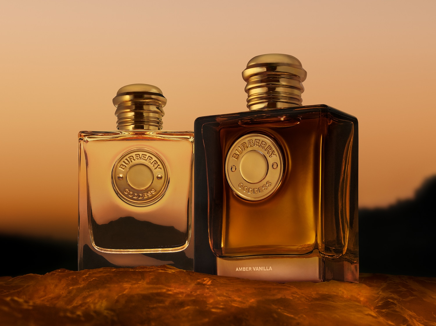 Deux flacons de parfum Burberry Goddess, l'un avec un liquide ambré clair et l'autre avec un liquide ambré plus foncé, reposent sur une surface ondulée brun-orange devant un ciel de coucher de soleil.