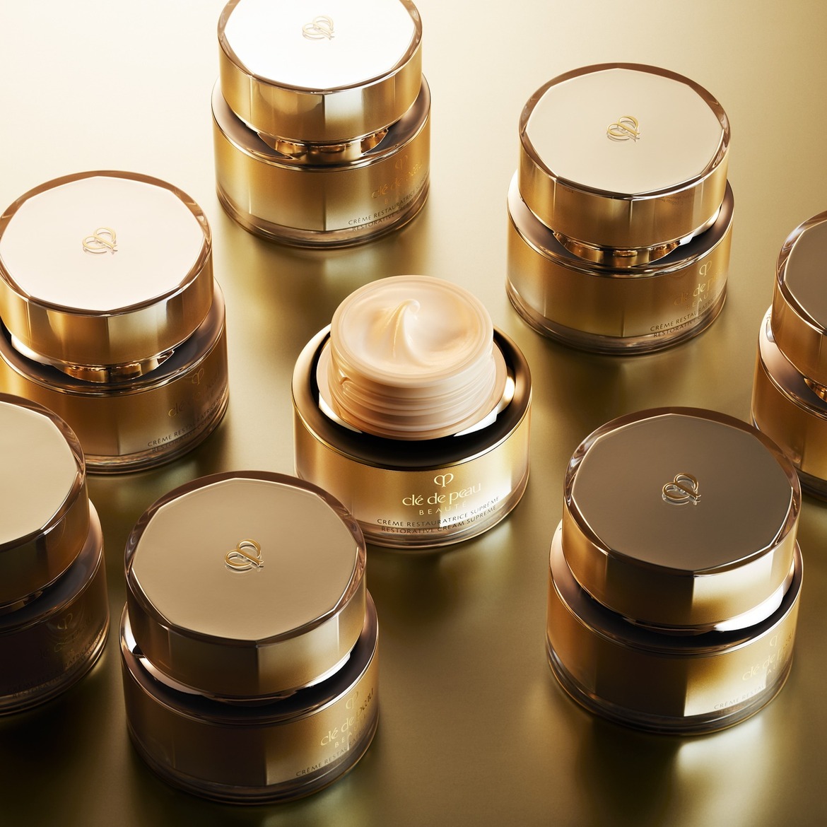 Een close-up van meerdere gouden potjes Clé de Peau Beauté Crème Restauratrice Suprême, waarvan één open is en de rijke, crèmekleurige textuur van de crème toont.