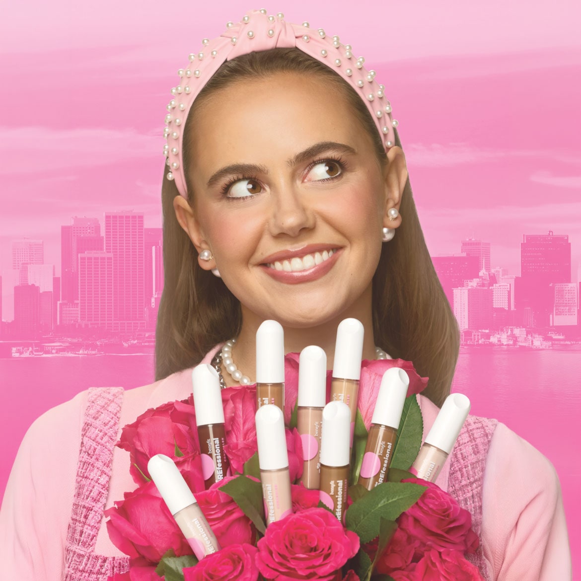 Een lachende vrouw met een roze haarband met parels en een roze jasje houdt een boeket rode rozen vast, waarin diverse Benefit Professional concealers in verschillende tinten zijn gestoken, tegen een roze achtergrond met een stadssilhouet.