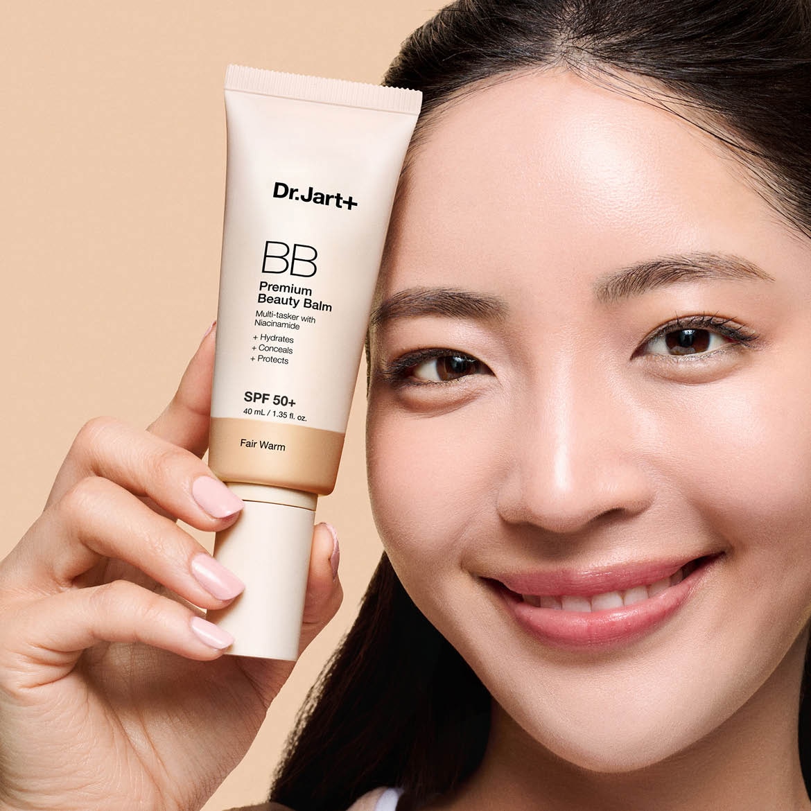 Une femme souriante tient un tube de Dr.Jart+ Premium Beauty Balm SPF 50+ dans la teinte Fair Warm, qui hydrate, camoufle et protège.