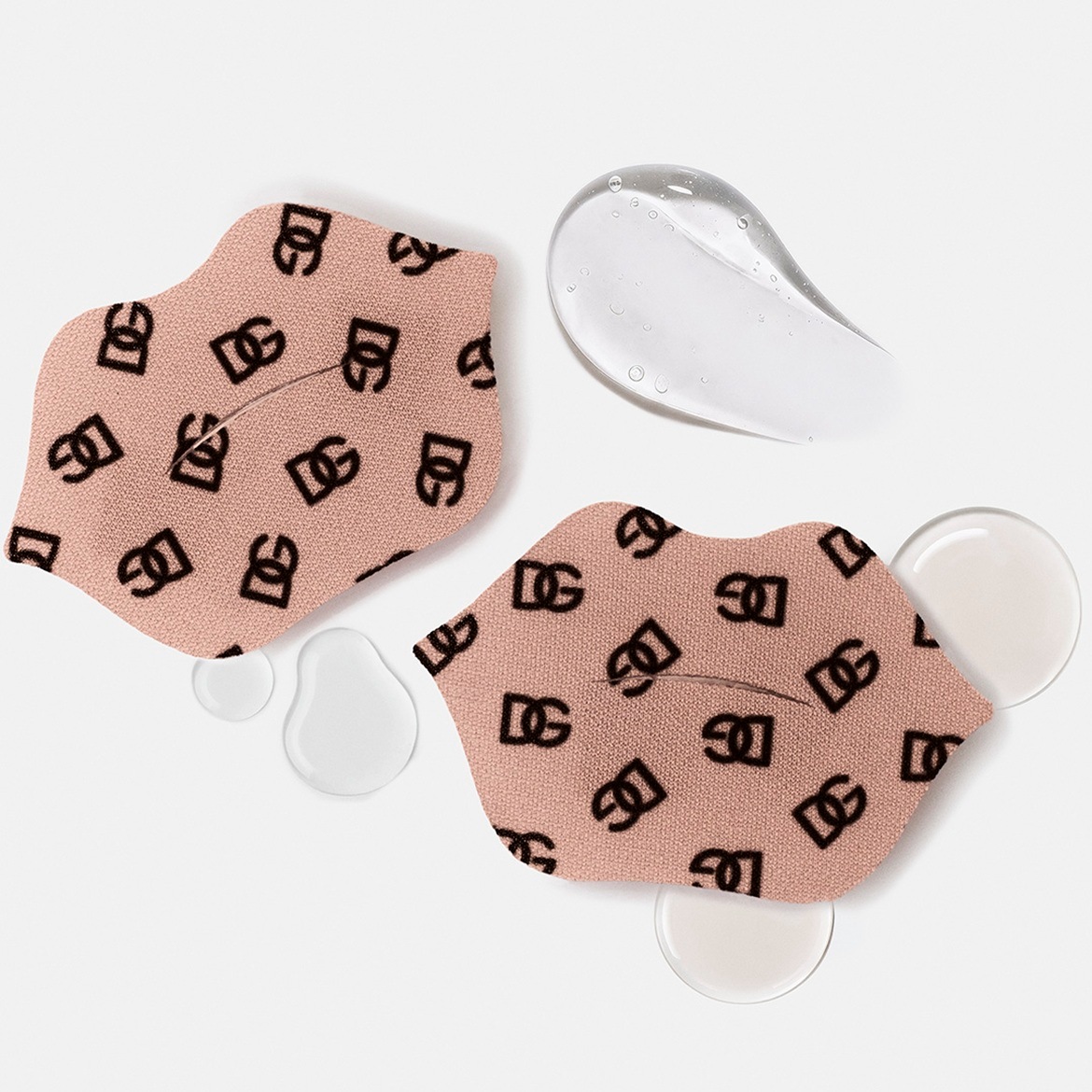 Deux masques pour les lèvres en forme de bouche, de couleur beige avec un motif répété du logo "DG" en noir, sont présentés avec des gouttes de liquide transparent et une texture de gel clair.