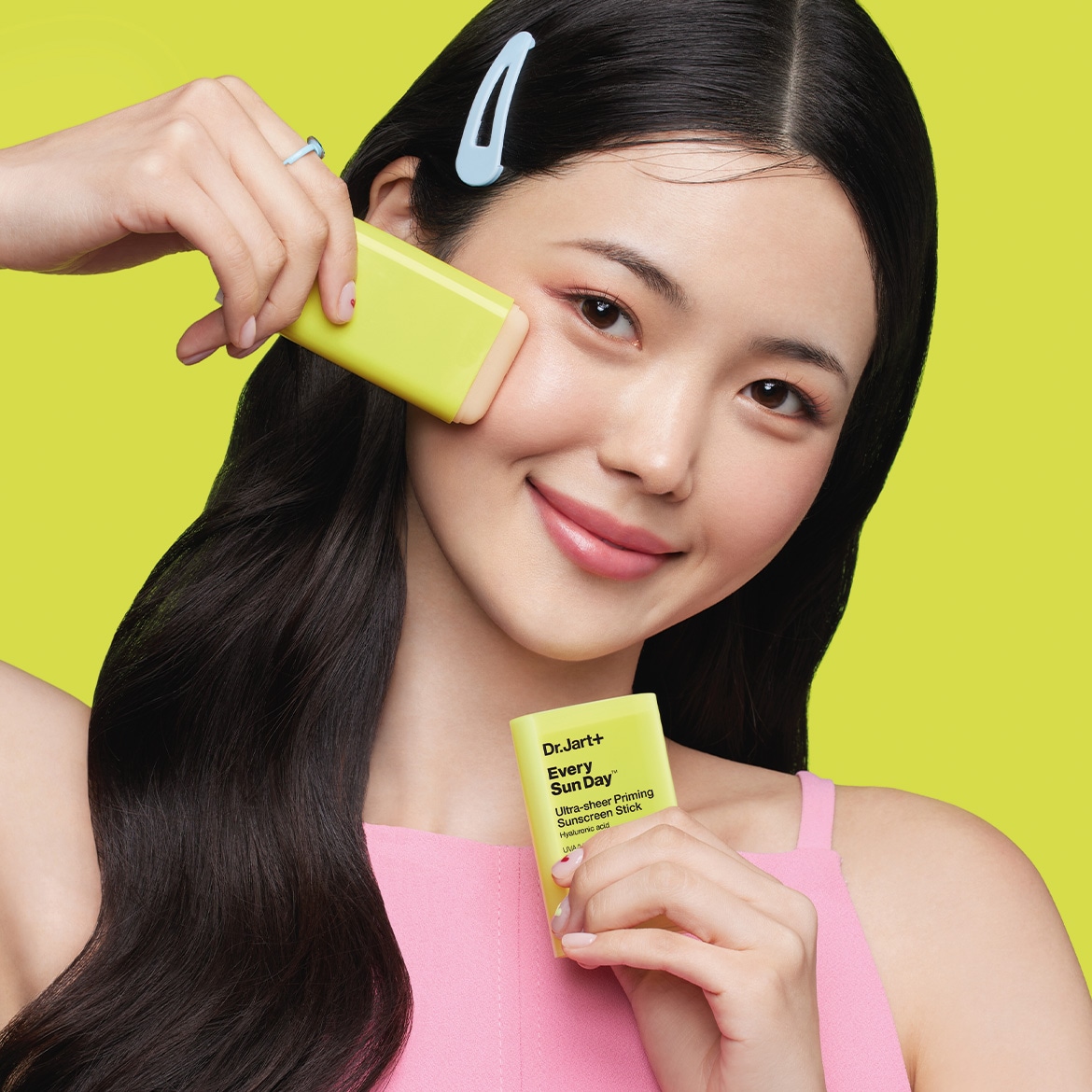 Une femme souriante applique le stick solaire Dr.Jart+ Every Sun Day™ sur sa joue, le packaging jaune vif promettant une protection solaire ultra-légère avec de l'acide hyaluronique.