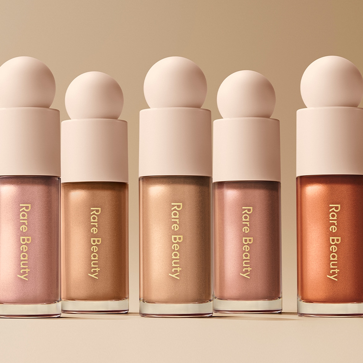 Vijf Rare Beauty vloeibare highlighters in verschillende tinten, van zachtroze tot diep brons, elk met een ronde dop, staan netjes op een rij.