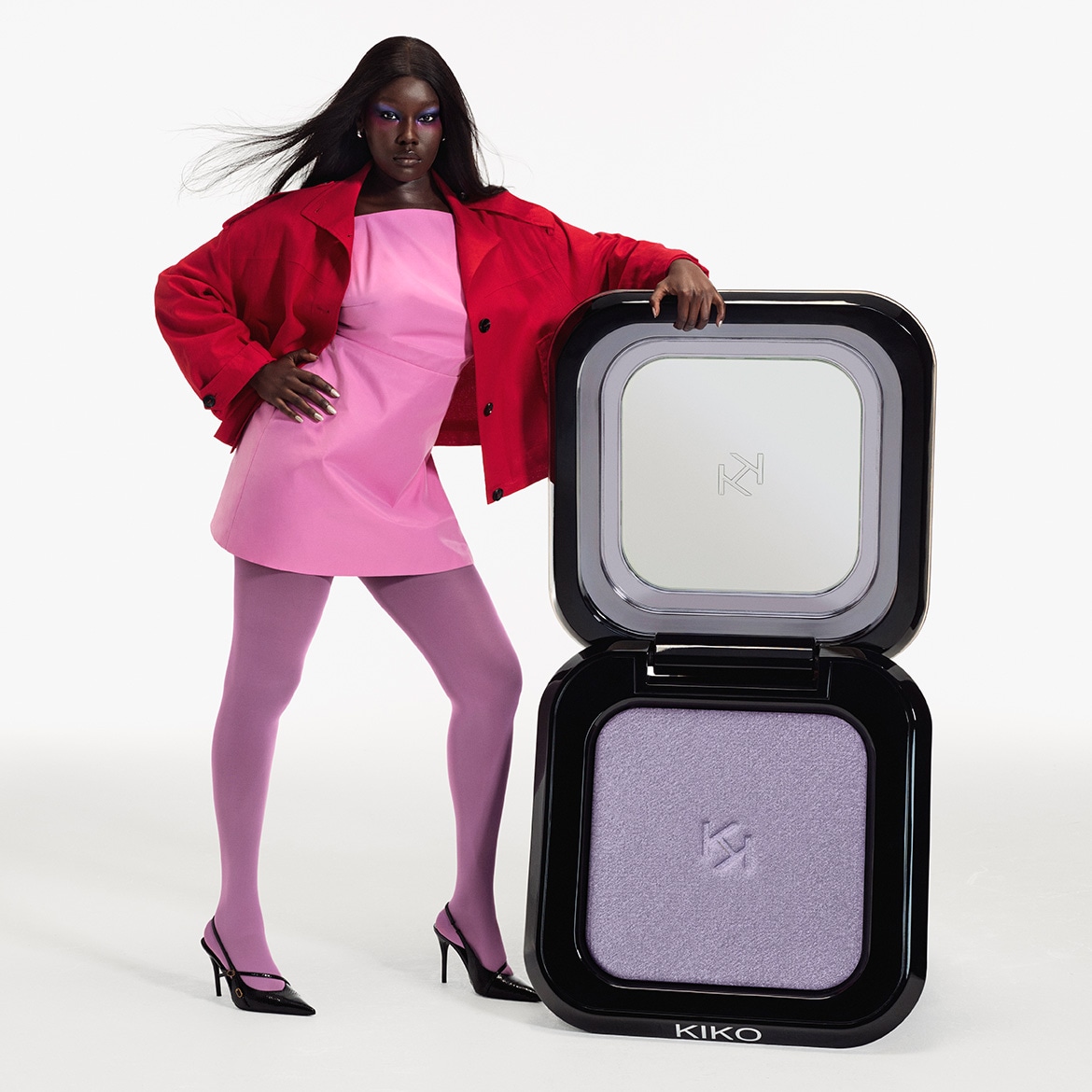 Un mannequin à la peau foncée, vêtue d'une robe rose, d'une veste rouge et de collants violets, pose à côté d'un poudrier compact ouvert de Kiko contenant un fard à paupières lilas.