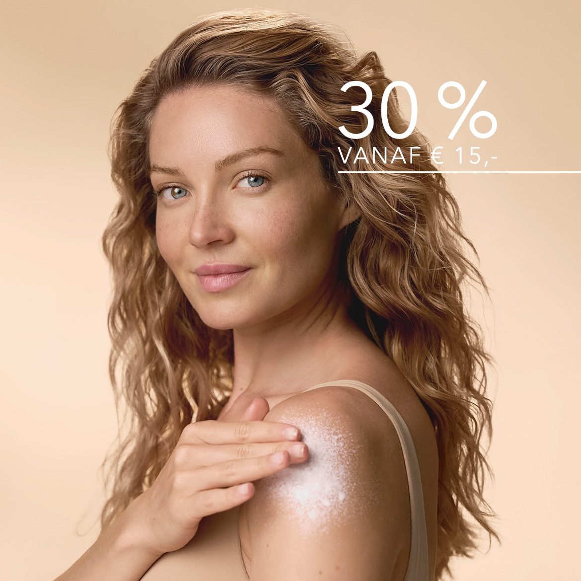 Een vrouw met krullend blond haar en een lichte huidskleur lacht terwijl ze een glinsterende lotion op haar schouder aanbrengt, met de tekst "30% VANAF € 15,-" in de rechterbovenhoek.