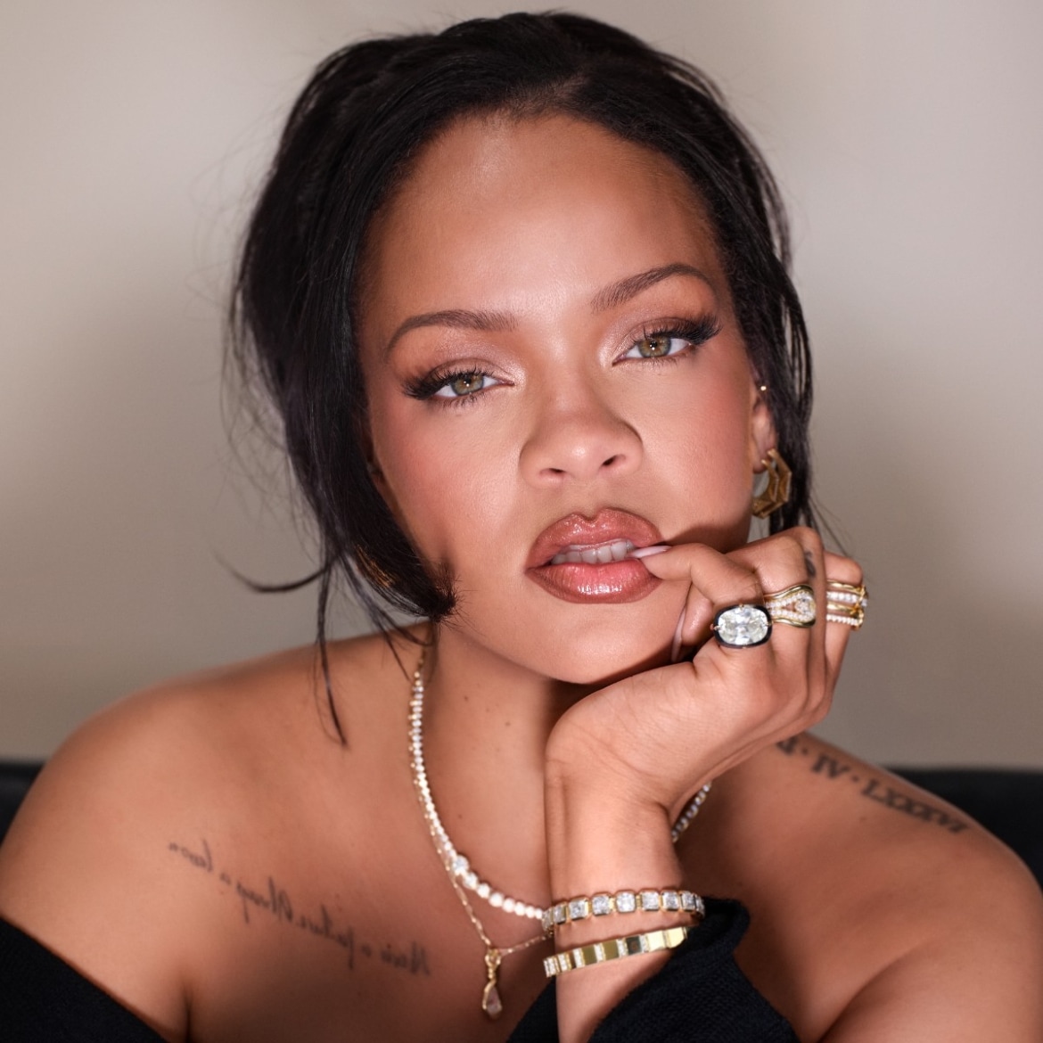 Rihanna porte un haut noir à épaules dénudées, ses cheveux foncés coiffés lâchement en arrière, et sa main ornée de plusieurs bagues repose sur son menton, tandis qu'elle se mordille légèrement les lèvres.