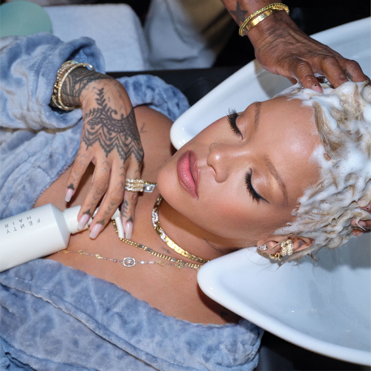 Rihanna est allongée et détendue dans un peignoir bleu clair, tandis que ses cheveux blonds sont lavés avec un shampoing riche et moussant dans un lavabo blanc, avec une bouteille blanche Fenty Hair à côté d'elle.