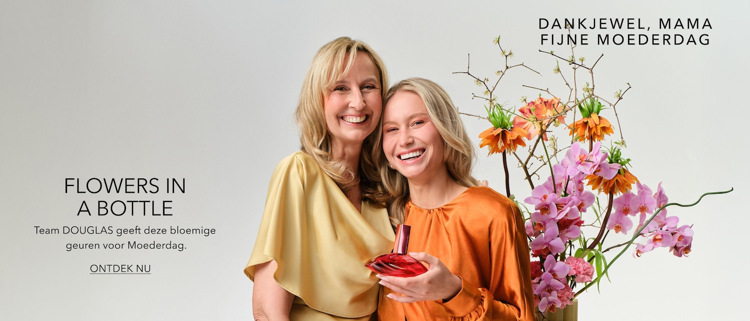 Twee lachende vrouwen, de ene in een gouden blouse en de andere in een oranje blouse, houden een rode parfumfles vast, omringd door een bloemstuk met roze en oranje bloemen, met de tekst 'Flowers in a bottle'.