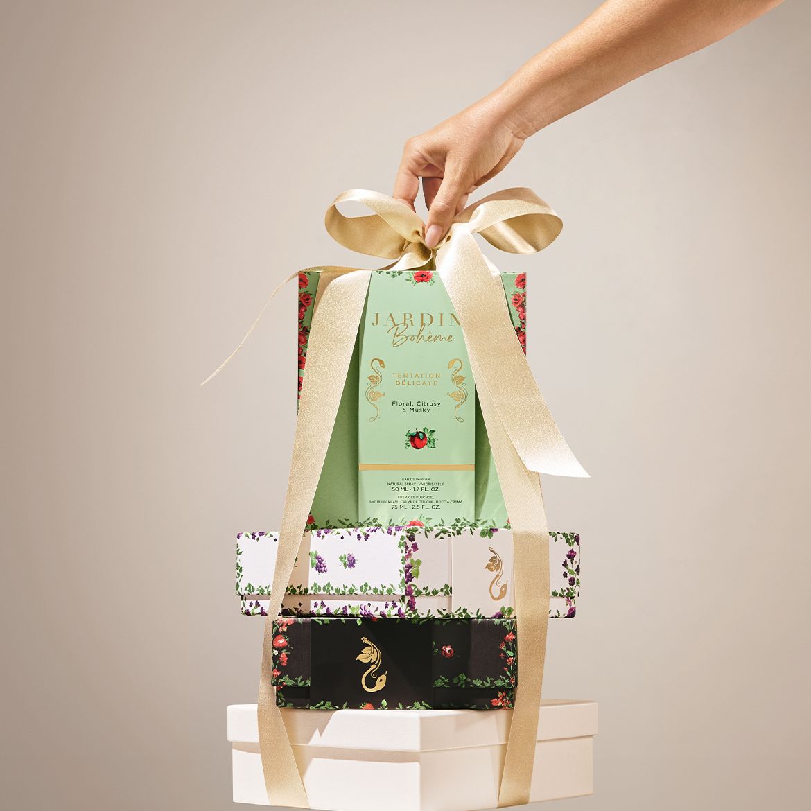 Een hand houdt een geschenkset van Jardin Bohème 'Tentation Délicate' vast, versierd met een gouden lint, met een parfum van 50 ml en een douchegel van 75 ml, gepresenteerd op een stapel decoratieve dozen.