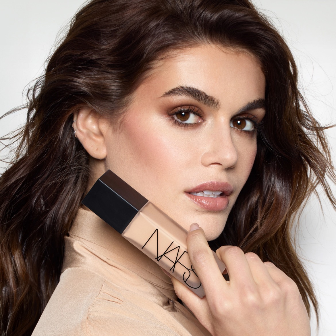 Een vrouw met bruin haar en ogen houdt een NARS foundation vast, de foundation heeft een zwarte dop en een doorzichtige fles.