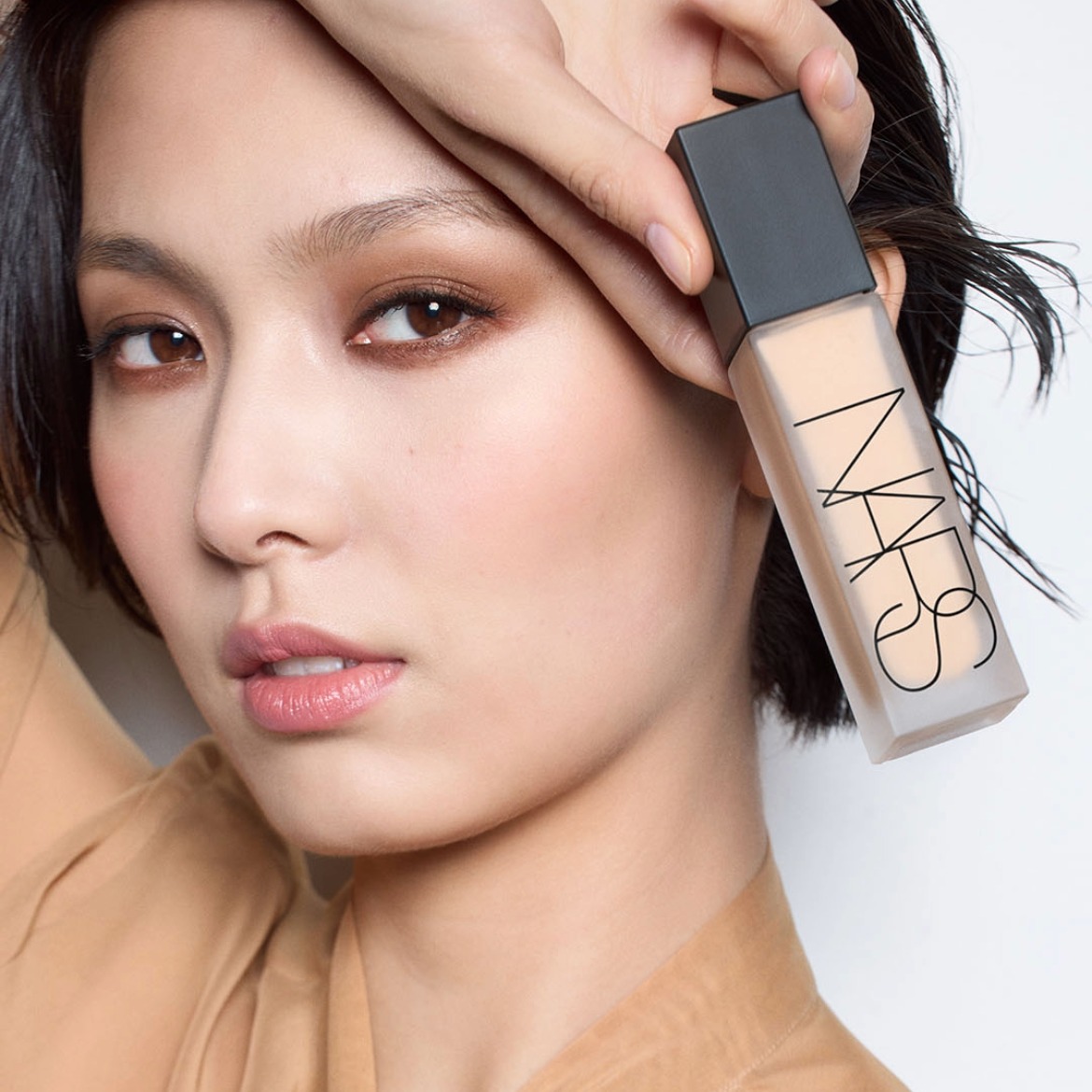 Een vrouw houdt een NARS foundationfles vast, met een matte dop en een doorschijnende fles die de zachte, beige tint van de foundation laat zien.