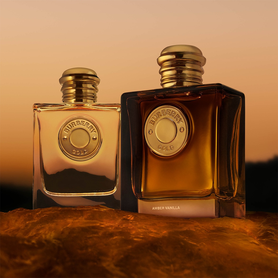 Deux parfums Burberry Gold Amber Vanilla dans des flacons carrés avec des bouchons et des étiquettes dorés, posés sur une surface brillante brun-orange devant un fond aux couleurs du coucher de soleil.