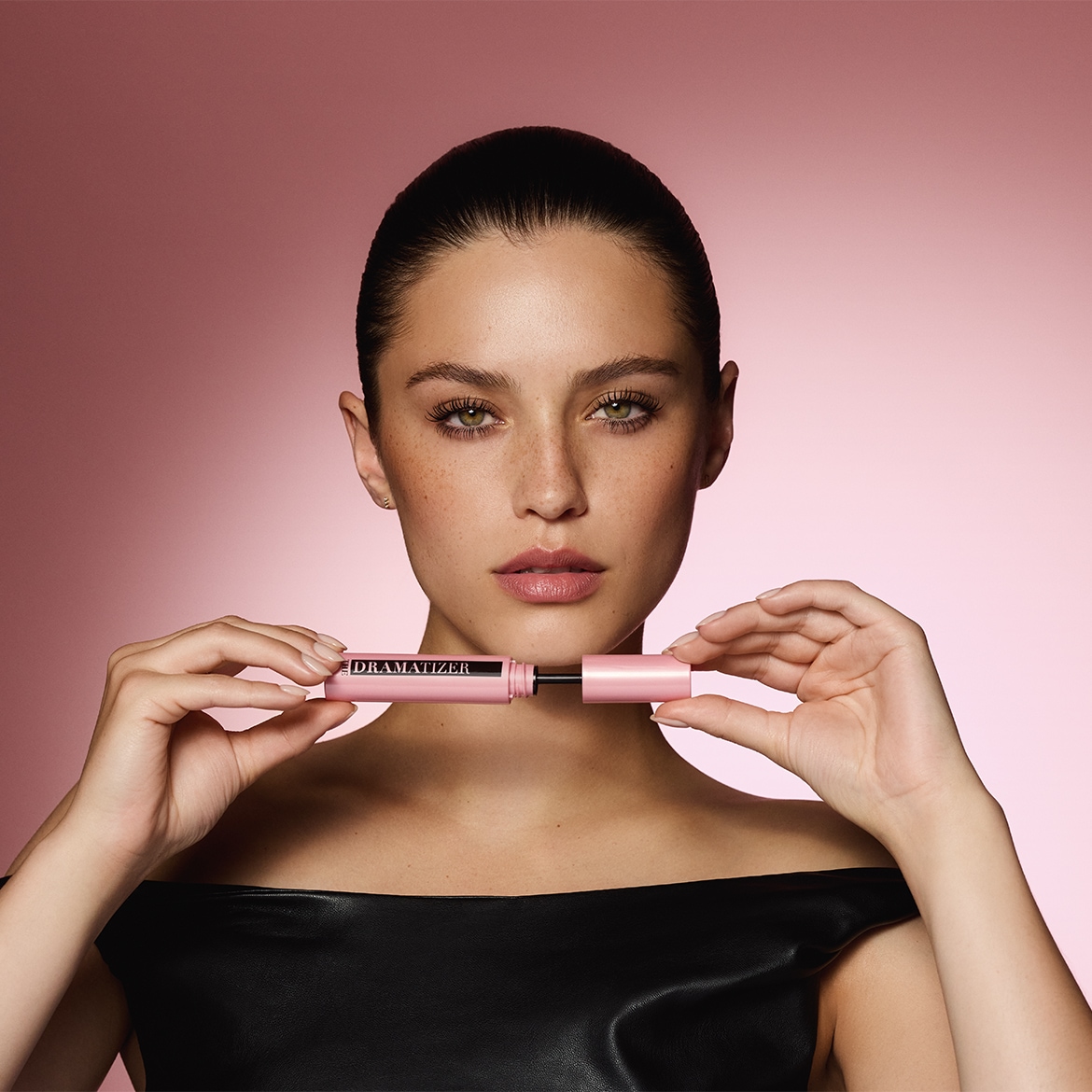 Een vrouw met een zwarte top houdt een roze Dramatizer mascara vast, klaar om je wimpers te transformeren met een dramatische look.