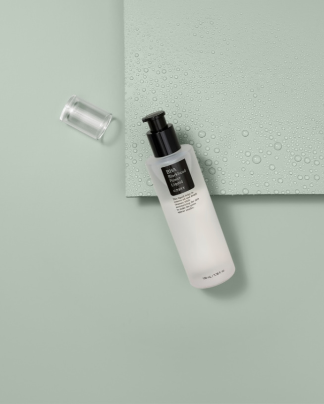 De Cosrx BHA Blackhead Power Liquid in een witte fles met een zwarte pomp ligt op een groene achtergrond met een transparante dop ernaast.