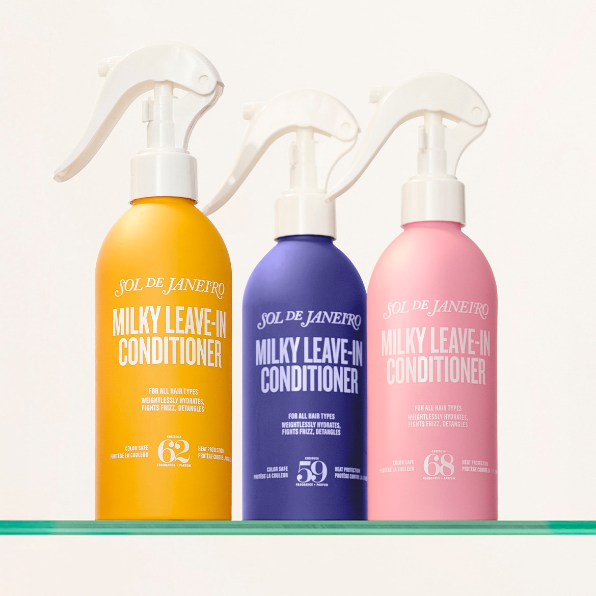 Trois revitalisants sans rinçage Sol de Janeiro Milky en jaune, violet et rose, chacun avec un vaporisateur blanc, sont posés sur une étagère en verre, offrant hydratation, lutte contre les frisottis et démêlage pour tous les types de cheveux.