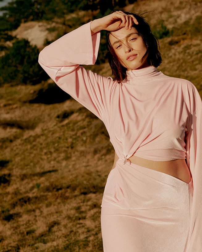 Een vrouw draagt een roze, nauwsluitende top met lange mouwen en een rok, staand in een veld met haar hand op haar voorhoofd.