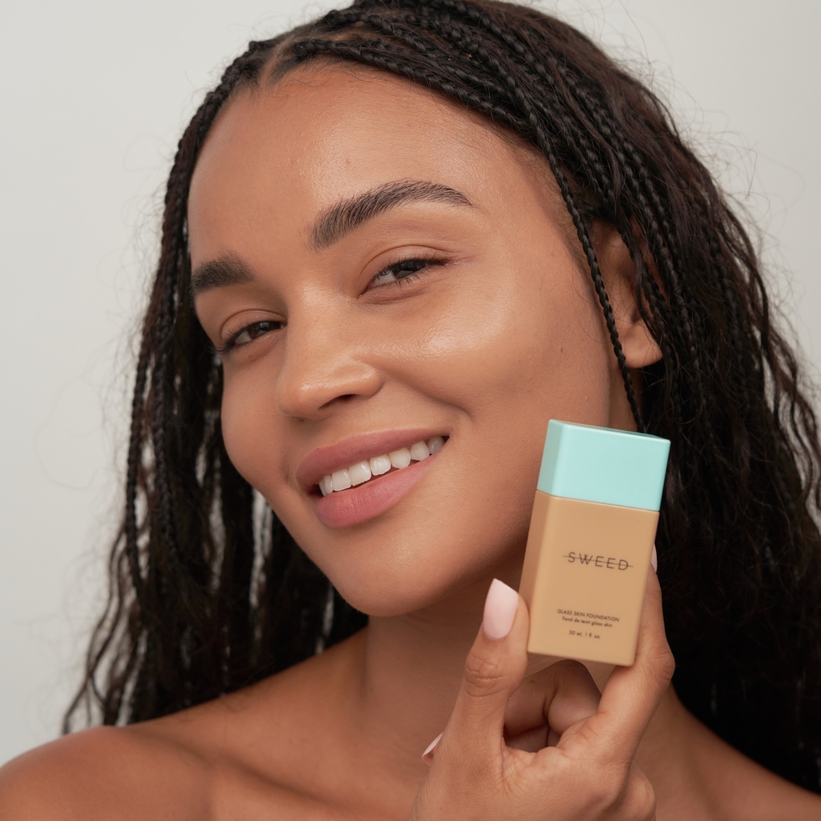 Une femme souriante tient un fond de teint SWEED au fini éclatant, avec un emballage beige et bleu clair, pour un teint impeccable.