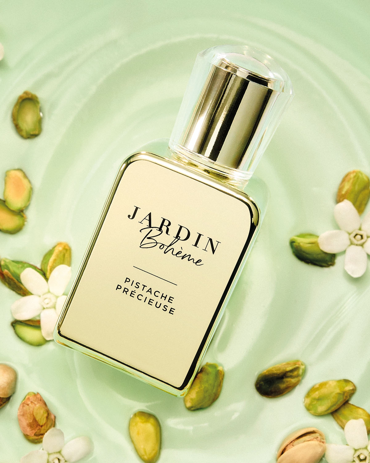 Un élégant flacon de parfum Jardin Bohème Pistache Précieuse, entouré de pistaches et de fleurs blanches sur un fond vert clair.