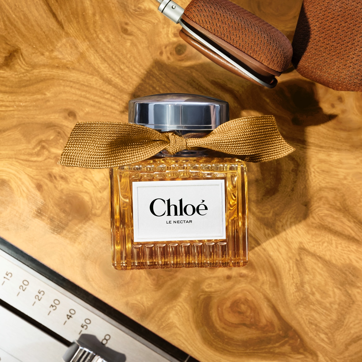 De Chloé Le Nectar parfumfles, versierd met een goudkleurige strik, staat op een houten ondergrond met een koptelefoon en een geluidsmixer op de achtergrond.