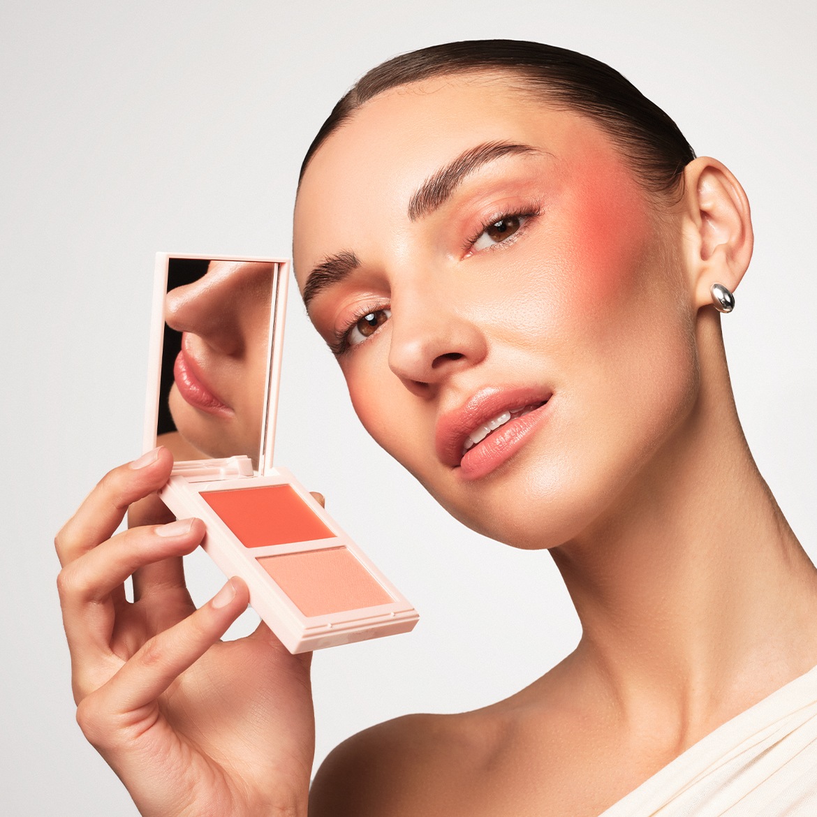 Une femme avec un fard à joues pêche sur les joues et les paupières tient un blush compact à deux teintes, l'une pêche et l'autre corail, avec un miroir reflétant ses lèvres et son nez.