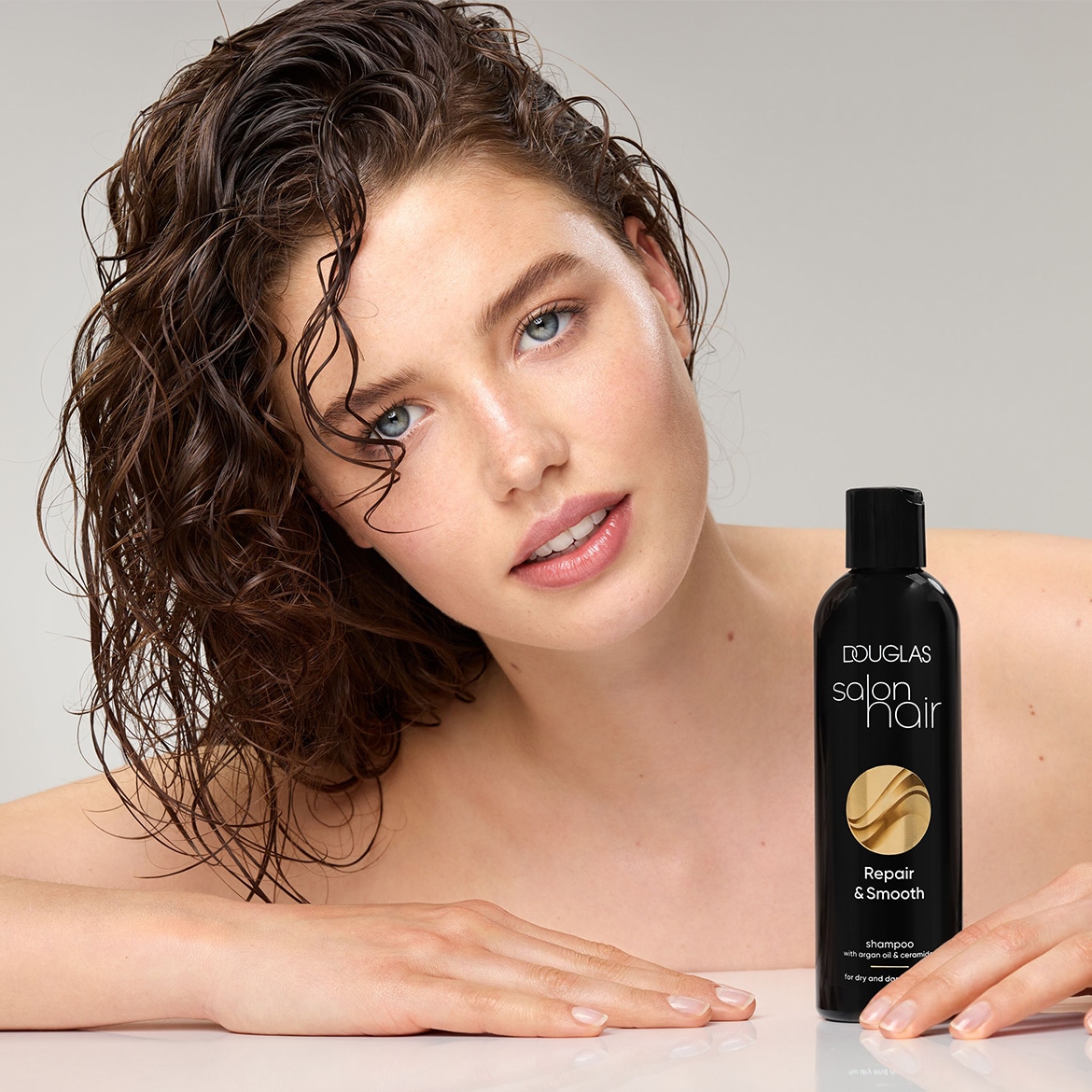 Een vrouw met nat, golvend haar en een natuurlijke make-up look, houdt een zwarte fles Douglas Salon Hair Repair & Smooth shampoo vast, speciaal voor droog en beschadigd haar.