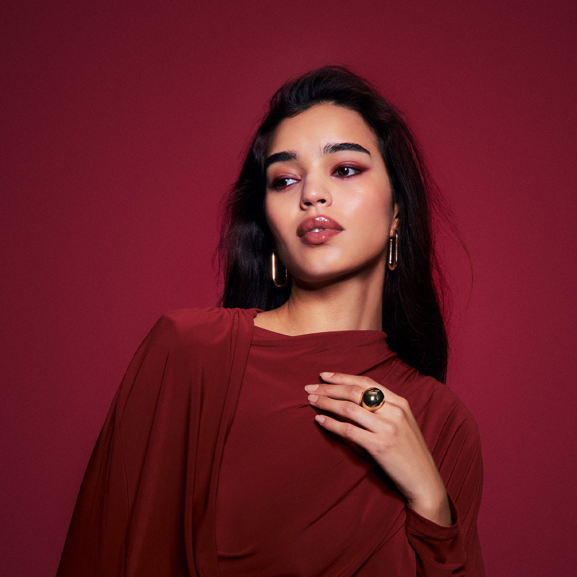 Een vrouw met donker haar, gouden oorbellen en een ring, gekleed in een bordeauxrode jurk, kijkt weg tegen een bordeauxrode achtergrond.
