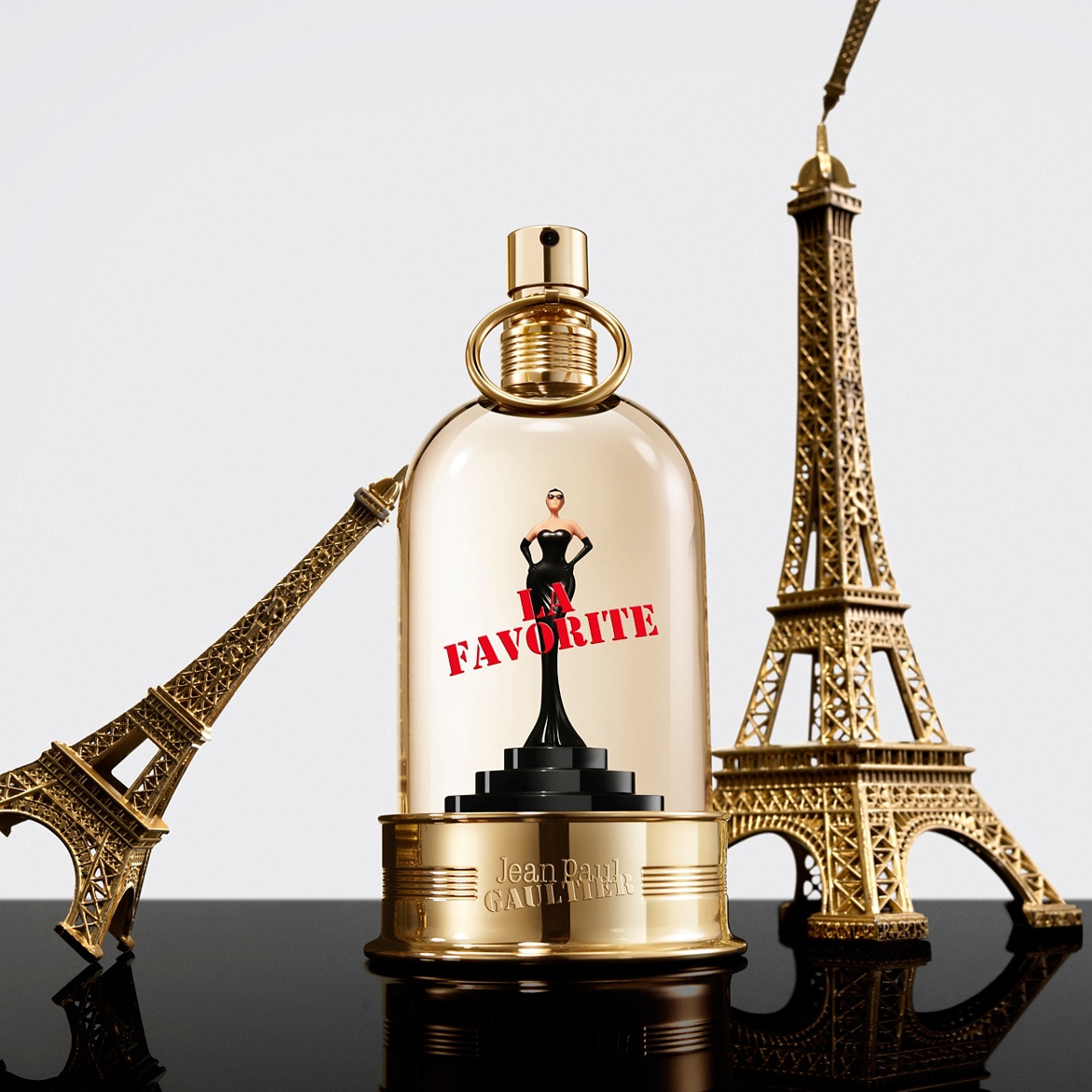 De Jean Paul Gaultier La Favorite parfumfles, met een gouden dop en een miniatuurbeeldje van een vrouw in een zwarte jurk, staat tussen twee gouden Eiffeltorenbeeldjes op een glanzend zwart oppervlak.